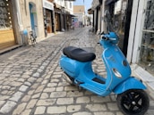 A bright blue Vespa scooter is parked on a cobblestone street. The area features small shops with glass displays and a bicycle propped against a wall. The street is narrow and lined with various storefronts, and a few people can be seen walking in the background.