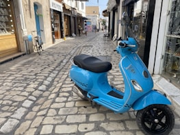 A bright blue Vespa scooter is parked on a cobblestone street. The area features small shops with glass displays and a bicycle propped against a wall. The street is narrow and lined with various storefronts, and a few people can be seen walking in the background.