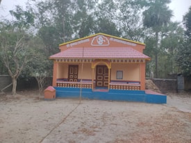 A small, colorful temple structure sits in a forested area. The building features orange walls with decorative blue accents and a red-tiled roof. An Om symbol is displayed above the main entrance, and there is textual signage in a regional script at the top. The area around the temple is sandy with scattered dry leaves, and trees surround the temple, giving it a serene and secluded feel.