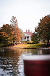 A serene landscape featuring a large, elegant house with a red roof, situated among tall, autumn-colored trees. The house is positioned on a grassy hill overlooking a calm body of water, reflecting the trees and the structure. The atmosphere is peaceful and picturesque.