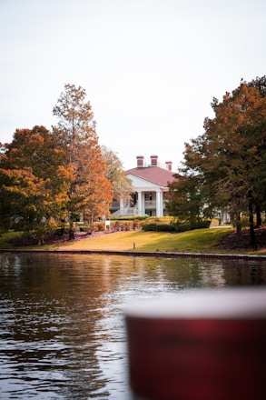 A serene landscape featuring a large, elegant house with a red roof, situated among tall, autumn-colored trees. The house is positioned on a grassy hill overlooking a calm body of water, reflecting the trees and the structure. The atmosphere is peaceful and picturesque.