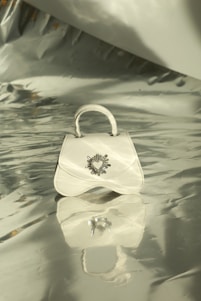 A small white handbag with a decorative heart emblem is centered on a shiny, reflective silver surface. Soft light and shadows play across the metallic background, creating an elegant and sophisticated setting.