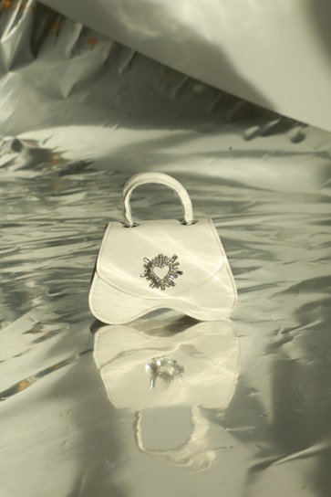 A small white handbag with a decorative heart emblem is centered on a shiny, reflective silver surface. Soft light and shadows play across the metallic background, creating an elegant and sophisticated setting.
