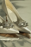 A pair of elegant high-heeled shoes made of satin material with sparkling crystal embellishments on the front. The shoes are positioned in a way that highlights their pointed toes and sleek design. The lighting adds a soft, luxurious glow to the image.