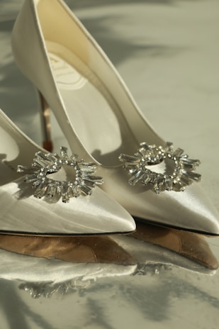 A pair of elegant high-heeled shoes made of satin material with sparkling crystal embellishments on the front. The shoes are positioned in a way that highlights their pointed toes and sleek design. The lighting adds a soft, luxurious glow to the image.