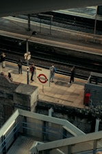 An elevated view of a train platform with a few people standing and waiting. The platform sign reads Paddington. Staircases and railings are visible in the foreground, with tracks and another platform in the background. The scene conveys the urban atmosphere of a train station.
