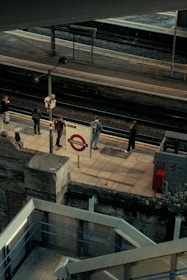 An elevated view of a train platform with a few people standing and waiting. The platform sign reads Paddington. Staircases and railings are visible in the foreground, with tracks and another platform in the background. The scene conveys the urban atmosphere of a train station.