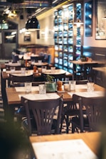 A cozy restaurant interior with several wooden tables and chairs neatly arranged, adorned with small decorative plants and condiments. The ambient lighting casts a warm glow, and shelves display various wine bottles along one wall. Large pendant lights hang from the ceiling, and the overall setting is clean and inviting.