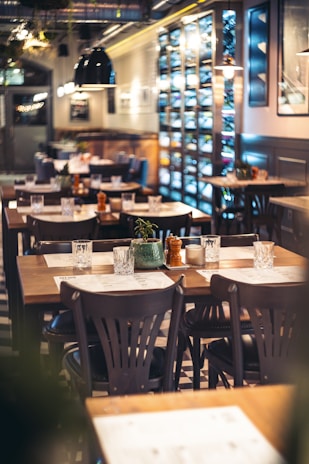 A cozy restaurant interior with several wooden tables and chairs neatly arranged, adorned with small decorative plants and condiments. The ambient lighting casts a warm glow, and shelves display various wine bottles along one wall. Large pendant lights hang from the ceiling, and the overall setting is clean and inviting.