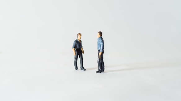 Two miniature figures are positioned facing each other on a plain white background. They appear to be in conversation, with both wearing dark clothing. The setting is minimalist, creating a sense of isolation or focus on the interaction between the figures.