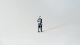 A small figurine of a human stands against a plain, light background. The figurine appears to be male and is wearing a blue top and black pants. It is positioned slightly off-center, creating a sense of space and minimalism.