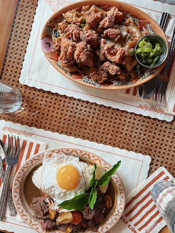 Two plates of appetizing food placed on a woven placemat. The top plate features fried chicken pieces on noodles with vegetables accompanied by a small bowl of sliced green chili. The bottom plate contains a fried egg over a bed of seasoned beef, cherry tomatoes, and fresh herbs. Both dishes are placed on decorative plates with cutlery on striped napkins.