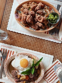 Two plates of appetizing food placed on a woven placemat. The top plate features fried chicken pieces on noodles with vegetables accompanied by a small bowl of sliced green chili. The bottom plate contains a fried egg over a bed of seasoned beef, cherry tomatoes, and fresh herbs. Both dishes are placed on decorative plates with cutlery on striped napkins.