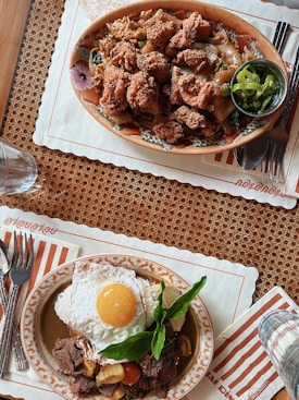 Two plates of appetizing food placed on a woven placemat. The top plate features fried chicken pieces on noodles with vegetables accompanied by a small bowl of sliced green chili. The bottom plate contains a fried egg over a bed of seasoned beef, cherry tomatoes, and fresh herbs. Both dishes are placed on decorative plates with cutlery on striped napkins.