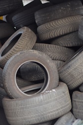 A pile of used tires is stacked haphazardly, with varying degrees of wear visible on the treads. The scene suggests an industrial setting or a recycling center, indicating the reuse or disposal of automotive parts.