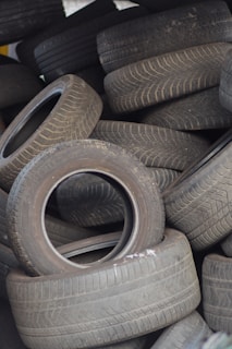 A pile of used tires is stacked haphazardly, with varying degrees of wear visible on the treads. The scene suggests an industrial setting or a recycling center, indicating the reuse or disposal of automotive parts.