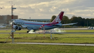 A large cargo airplane with markings indicating it's operated by Cargolux is landing on an airport runway. Smoke is visible near the landing gear, suggesting the moment of touchdown. The surroundings include grassy fields, trees, and some airport facilities in the background under a partly cloudy sky.