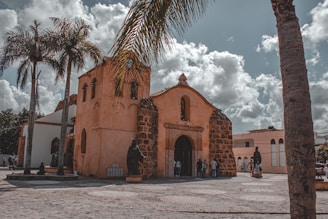A historic church with a stone facade and arched entrance is surrounded by tall palm trees under a partly cloudy sky. Several people stand near the entrance and statues are visible in the courtyard.