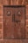 A wooden door with two round stained glass windows featuring crosses and intricate designs. The door has a rustic, weathered look with visible patina. There are two metal knockers and a large padlock securing the door. Surrounding the door is a brick wall with a rough texture.