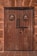 A wooden door with two round stained glass windows featuring crosses and intricate designs. The door has a rustic, weathered look with visible patina. There are two metal knockers and a large padlock securing the door. Surrounding the door is a brick wall with a rough texture.