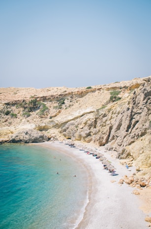A serene beach scene featuring clear turquoise waters meeting a pebbly shore. Sunbathers and swimmers can be seen along the coastline. Sun umbrellas and a few chairs are set up on the beach. The backdrop features rocky cliffs with sparse vegetation under a clear blue sky.