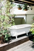 A white freestanding bathtub is situated outdoors on a wooden platform surrounded by lush green plants. Hanging fern baskets are suspended from the ceiling, creating a natural and serene atmosphere under a covered area with ample sunlight.