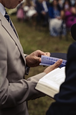 A person wearing a beige suit holds a program or booklet in one hand and a microphone in the other. In the background, a blurred group of people in formal attire can be seen sitting on chairs, suggesting a formal or ceremonial event.