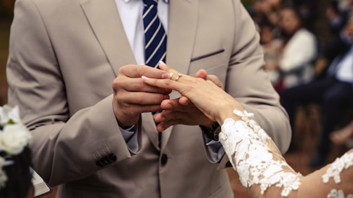 A groom places a wedding ring on the bride's finger, both dressed in formal attire. The groom is wearing a beige suit and a striped tie, while the bride has a detailed lace sleeve. The background is blurred, showing people sitting.