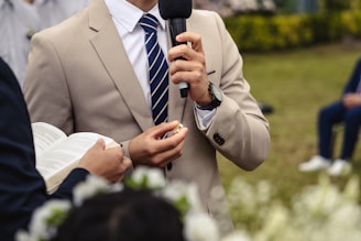 A person in a beige suit and striped tie holding a microphone, with another person holding an open book nearby. The event appears formal, taking place outdoors on a grassy area. There are blurred flowers in the foreground and a seated person in the background.