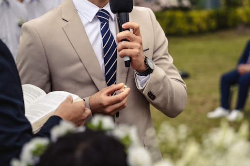 A person in a beige suit and striped tie holding a microphone, with another person holding an open book nearby. The event appears formal, taking place outdoors on a grassy area. There are blurred flowers in the foreground and a seated person in the background.