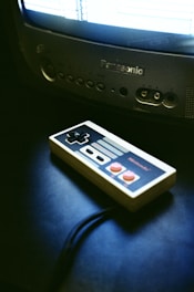 A vintage game controller rests on a dark surface next to an old CRT television. The television is turned on, displaying a bright screen with indistinct graphics. The atmosphere suggests a retro gaming setup.