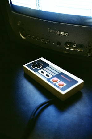 A vintage game controller rests on a dark surface next to an old CRT television. The television is turned on, displaying a bright screen with indistinct graphics. The atmosphere suggests a retro gaming setup.