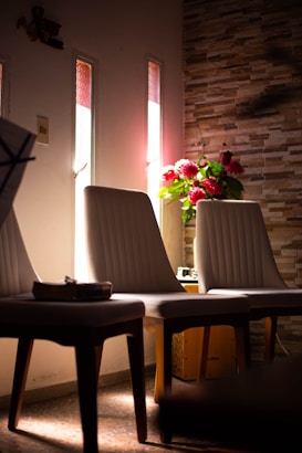 A warmly lit room features two cushioned chairs positioned against a wall with a textured stone-like design. Sunlight filters through vertical windows, casting soft shadows across the space. A decorative vase with vibrant red flowers and lush green leaves adds a touch of color beside the chairs.
