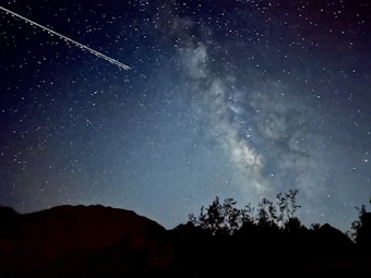 A stunning night sky filled with countless stars and a prominent streak of light, possibly from a satellite or meteor, over a silhouetted landscape of trees and hills. The Milky Way galaxy is visible, creating a celestial pathway across the dark expanse.