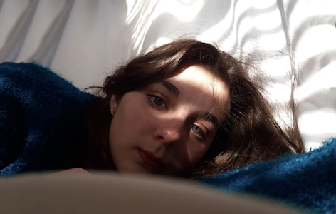 A woman is lying down with her head resting on a dark blue blanket, her face partially illuminated by a soft, dappled light. Her eyes are focused, and her hair is slightly tousled, blending into the blanket.