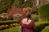 A person with a braided hairstyle in a patterned dress stands in a manicured garden surrounded by various types of lush greenery and colorful shrubs.