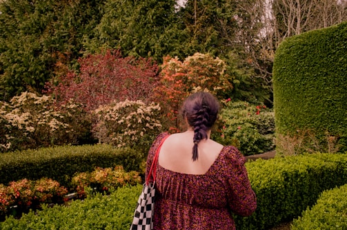 A person with a braided hairstyle in a patterned dress stands in a manicured garden surrounded by various types of lush greenery and colorful shrubs.