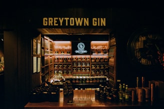 A dimly lit gin store displays a wide variety of bottles organized on shelves. The central focus is a backlit sign reading 'GREYTOWN GIN' above the shelves. A counter in the foreground contains bottle packs and flyers, while a large wall clock decorates one side.