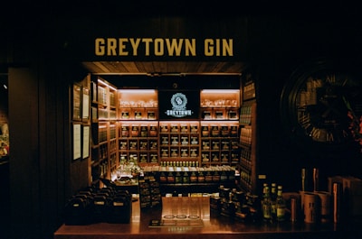A dimly lit gin store displays a wide variety of bottles organized on shelves. The central focus is a backlit sign reading 'GREYTOWN GIN' above the shelves. A counter in the foreground contains bottle packs and flyers, while a large wall clock decorates one side.
