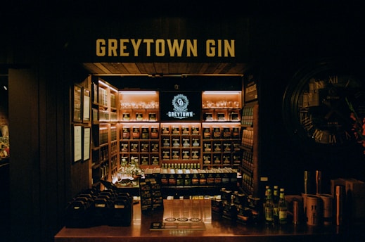 A dimly lit gin store displays a wide variety of bottles organized on shelves. The central focus is a backlit sign reading 'GREYTOWN GIN' above the shelves. A counter in the foreground contains bottle packs and flyers, while a large wall clock decorates one side.