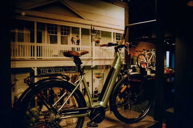 A vintage-style bicycle is prominently displayed indoors against a backdrop of a street scene painting featuring a building with a sign that reads 'The White Swan.' The background shows a couple of people standing on a balcony. The bicycle has a light green frame, brown leather saddle, and handlebars.
