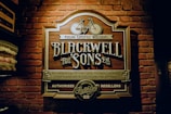 A vintage-style sign with ornate lettering is mounted on a brick wall. The sign displays 'Blackwell and Sons,' promoting cycling lifestyle merchants and emphasizing fine quality merchandise for outdoor recreation and gifting. A vintage bicycle graphic is featured at the top of the sign.
