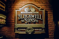 A vintage-style sign with ornate lettering is mounted on a brick wall. The sign displays 'Blackwell and Sons,' promoting cycling lifestyle merchants and emphasizing fine quality merchandise for outdoor recreation and gifting. A vintage bicycle graphic is featured at the top of the sign.