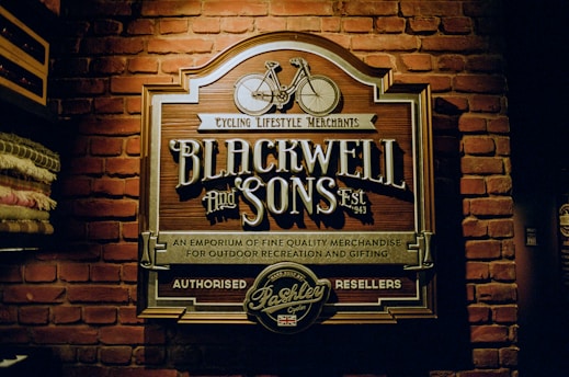 A vintage-style sign with ornate lettering is mounted on a brick wall. The sign displays 'Blackwell and Sons,' promoting cycling lifestyle merchants and emphasizing fine quality merchandise for outdoor recreation and gifting. A vintage bicycle graphic is featured at the top of the sign.