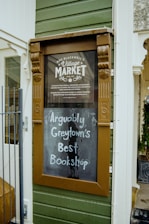 An outdoor sign with a vintage wooden frame is mounted on the exterior green wooden paneling of a building. The top section of the sign reads 'Mrs Blackwell’s Village Market' with decorative elements, and the lower portion features handwritten text on a blackboard stating, 'Arguably Greytown’s Best Bookshop.'