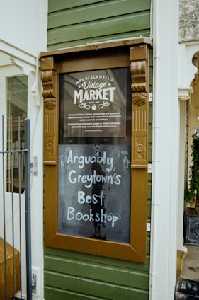An outdoor sign with a vintage wooden frame is mounted on the exterior green wooden paneling of a building. The top section of the sign reads 'Mrs Blackwell’s Village Market' with decorative elements, and the lower portion features handwritten text on a blackboard stating, 'Arguably Greytown’s Best Bookshop.'