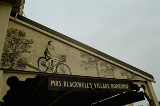 A mural on the facade of a building features an illustration of a person riding a bicycle, with a basket attached, surrounded by trees. Below the mural, there is a sign that reads 'MRS BLACKWELL'S VILLAGE BOOKSHOP.'