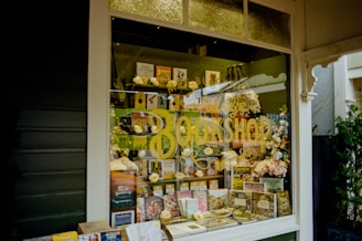 A cozy bookshop window display with a variety of books and floral decorations. The window features yellow text reading 'BOOKSHOP' and is filled with colorful books of different genres, some placed upright, others stacked. Decorative flowers and small gift items are arranged around the books, adding a welcoming and quaint charm to the display.