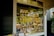A cozy bookshop window display with a variety of books and floral decorations. The window features yellow text reading 'BOOKSHOP' and is filled with colorful books of different genres, some placed upright, others stacked. Decorative flowers and small gift items are arranged around the books, adding a welcoming and quaint charm to the display.