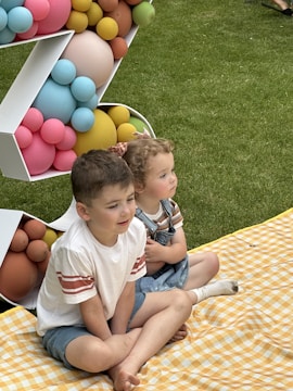 Two young children are sitting on a yellow checkered picnic blanket laid on grass. They are positioned beside a colorful balloon decoration consisting of pastel different-colored spheres. The children appear relaxed and are dressed in casual summer clothes.